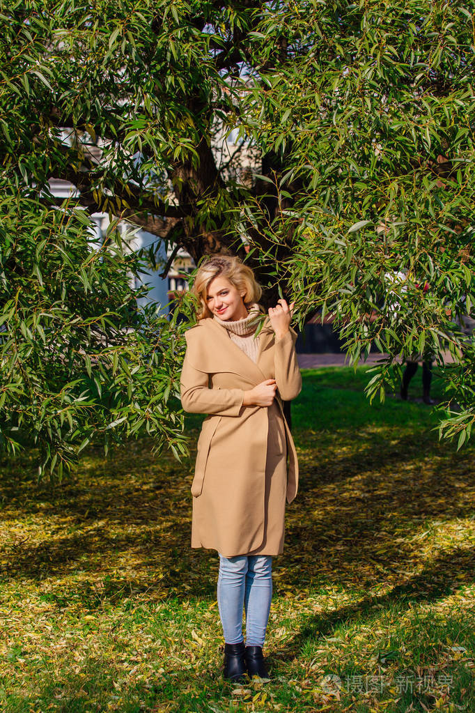 美丽优雅的金发女郎穿着大衣站在树下的秋天公园晴朗的10月天