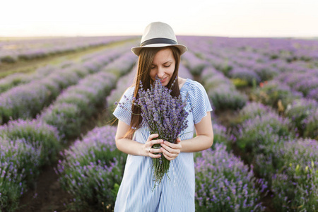 年轻的美女穿着蓝色的连衣裙, 帽子上紫色的薰衣草花草田野户外的夏天自然背景。嫩的雌性在开花灌木附近与花束。生活方式概念