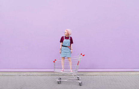 时尚的女孩粉红色的头发站在购物车的背景下, 一个紫色的墙。女孩时髦和购物车