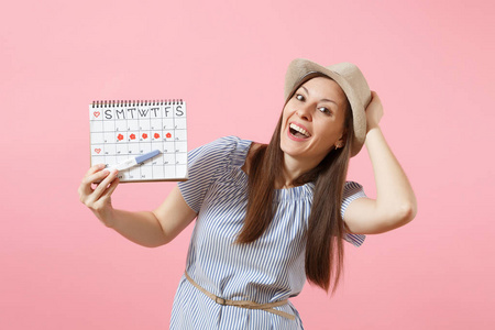 兴奋的快乐女人穿着蓝色的连衣裙, 戴着帽子手里的怀孕测试, 月经期的日历用来检查在粉红色背景下被隔离的经期。医疗保健妇科概念复制