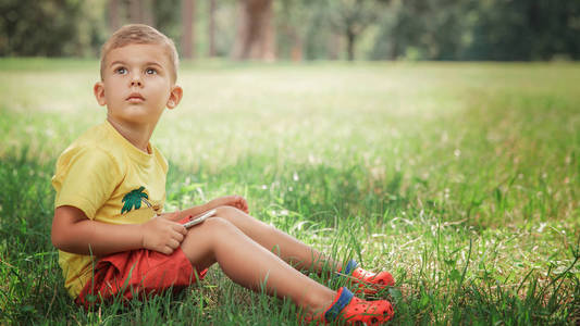 一个小男孩正坐在草地上的电话里. 穿着黄色 t恤和红色短裤的小男孩坐在草地上用智能手机