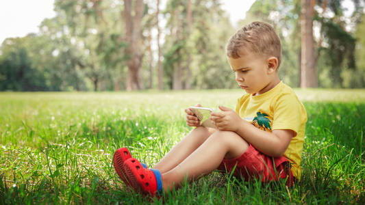 一个小男孩正坐在草地上的电话里. 穿着黄色 t恤和红色短裤的小男孩坐在草地上用智能手机
