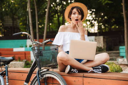 在公园里坐着膝上型电脑的年轻女孩穿着夏天的衣服