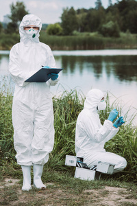 女科学家在保护面具和西装写在剪贴板, 而她的同事把水样品附近的河流室外
