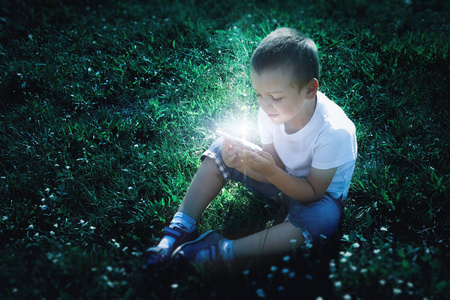 男孩坐在草地上, 手里拿着一把神奇的手机。黑暗背景下的魔法发光, 光线和光辉的阴影