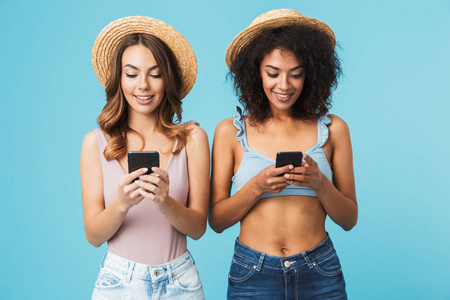 两个合群的多民族妇女20s 戴草帽微笑, 并使用手机在蓝色背景下隔离