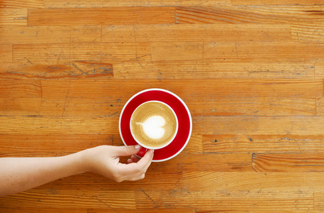 年轻女子手捧红杯咖啡与心形泡沫拿铁艺术的最高视图。女性喝卡布奇诺, 坐在 grunged 划伤的木条桌上。背景, 特写, 复制空