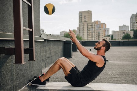 英俊的运动员侧面观看在屋顶上的瑜伽垫与医学球坐在 ups 上