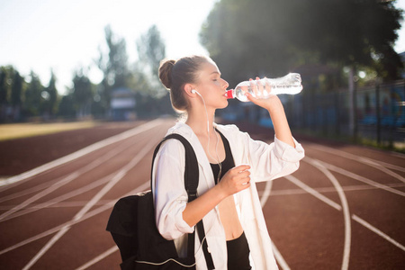 美丽的女孩在运动穿着背包在肩上饮用纯净水在体育场跑道上