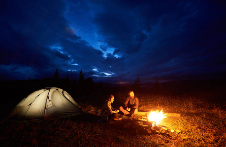 年轻夫妇的男人和女人在夜间露营在山上享受, 坐在燃烧篝火附近, 并照亮了美丽的傍晚多云天空的旅游帐篷。旅游理念