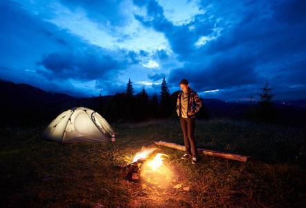 年轻女子徒步旅行者晚上在山上露营, 坐在燃烧的篝火旁, 在美丽的傍晚多云的天空下照亮了旅游帐篷。旅游户外活动理念