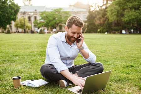 成人欧洲男子的照片在商业服装坐在草地上的腿交叉, 并在手机上发言, 而在银色笔记本电脑工作