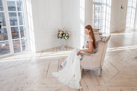 穿着白色礼服的漂亮金发女郎的时尚照片坐在扶手椅上反对白色内饰背景