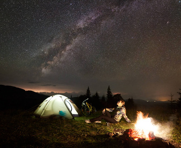 年轻女子游客在夜间露营, 在燃烧的篝火旁野营, 照亮旅游帐篷, 山地自行车在令人难以置信的美丽的夜空充满星星和银河。旅游理念