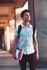 学生拿着笔记本, 背着背包走在大学大楼的前面。穿着蓝色衬衣和深色牛仔裤的年轻男孩