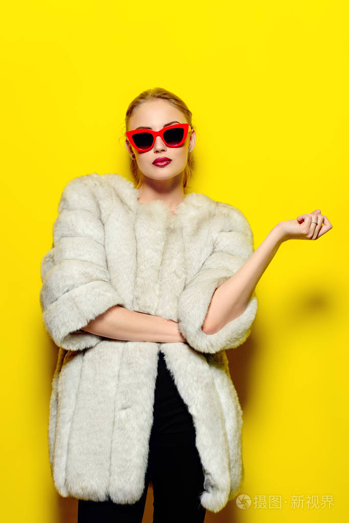 华丽的金发女人摆在奢华的毛皮大衣和别针太阳镜.黄色背景.