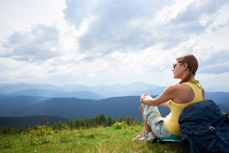 后视图的快乐女子背包坐在草地上的山上休息, 带着背包, 戴着太阳镜。在山中享受夏日多云天气的女性旅行者。户外活动, 旅游理念