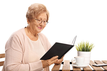 老妇人坐在茶几边看书, 看着一本白色背景的书。