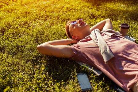在春天的校园公园里, 英俊的大学男子不寒而栗。快乐的家伙学生躺在草地上, 他的电话和咖啡旁边的日落