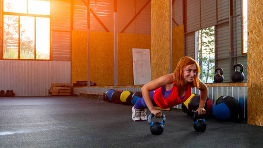 一个年轻的红头发的女人在粉红色的顶部和紫色的紧身裤做俯卧撑, 手在体育健身房的重量。重推技术