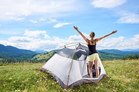 年轻女子观光徒步登山小径, 站在帐篷内, 举起手在空中, 享受夏日阳光明媚的早晨在山上。冒险假期户外概念