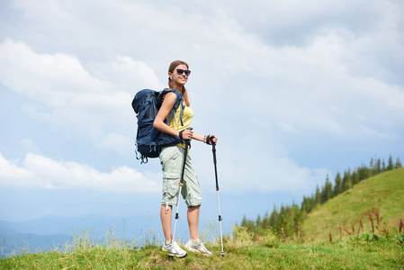 迷人的微笑的女子旅游徒步登山小径, 走在草地上, 穿着背包和太阳镜, 使用徒步棍, 享受夏季多云的一天。户外活动, 旅游理念