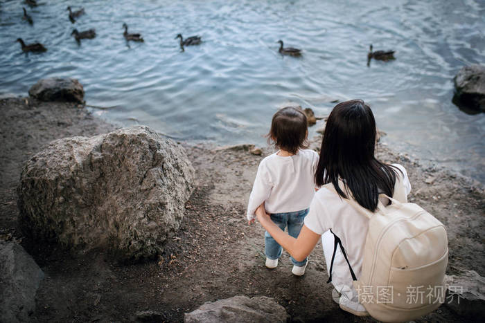 后视图的母亲和蹒跚学步的女孩坐在湖边鸭在河的背景.