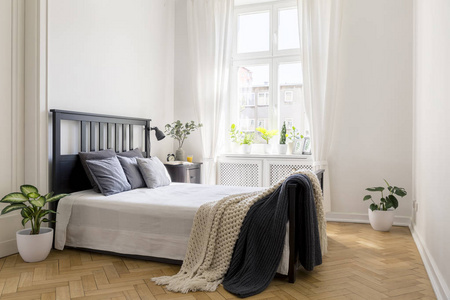 在床上的针织毯子在最小的白色卧室内部与植物和窗口。真实照片