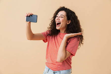 幸福年轻的卷发妇女的形象站在米色背景下, 使自拍用手机交谈