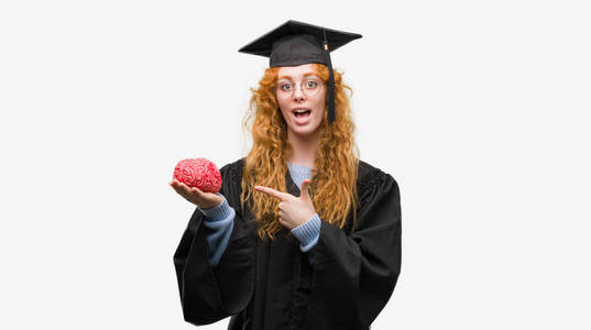 年轻红发学生女子穿着毕业制服持有的大脑非常高兴地指着手和手指