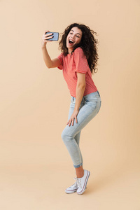 一个开朗的年轻女孩的全长肖像采取自拍与手机在米色背景下隔离