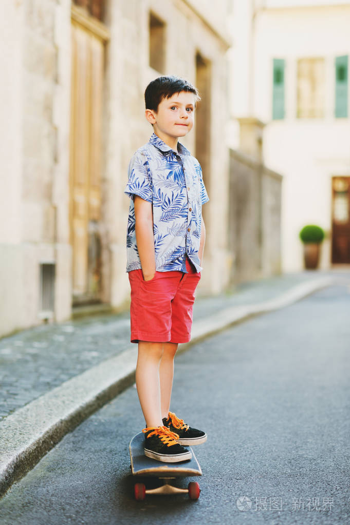 有趣时尚的孩子男孩摆了滑板在户外,穿着蓝色印花衬衫和红色短裤
