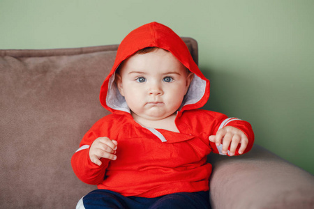 肖像可爱可爱的白种人微笑的小男孩蓝眼睛, 穿着红色运动衫帽衫和运动服裤。七月的孩子坐在沙发上在家里看着相机