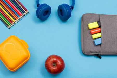 午餐盒, 彩色铅笔, 耳机, 苹果和袋铅笔盒的彩色毡笔和标记在蓝色背景。具有复制空间的顶部视图。回到学校的概念。学校用品