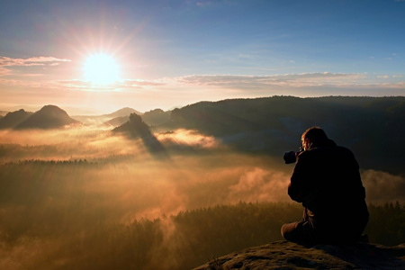 用手中的相机快乐旅游坐在砂岩岩石和观看到丰富多彩的雾，雾的早晨谷峰