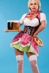 年轻慕尼黑啤酒节服务员，穿连衣裙的女孩传统巴伐利亚，服务在蓝色背景上的大啤酒杯