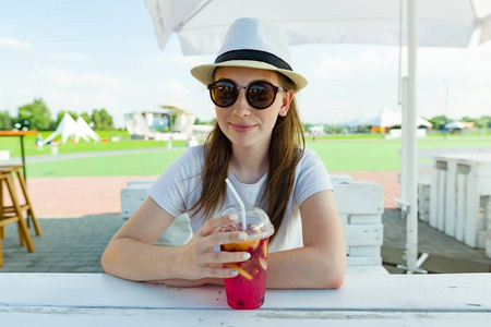 暑假休息吧16岁的少女在帽子和太阳镜与浆果饮料坐在桌子上夏季街咖啡馆。背景绿色休闲娱乐区