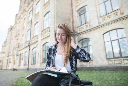 一个穿着衬衣的正面女孩坐在大学大楼附近的长凳上, 看书和微笑。有吸引力的女学生在大学校园里从事自我教育