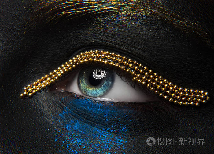 宏观和特写创意化妆主题: 美丽的女性眼睛黑皮肤与金链和蓝色颜料