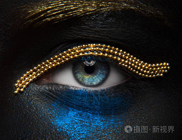 宏观和特写创意化妆主题: 美丽的女性眼睛黑皮肤与金链和蓝色颜料