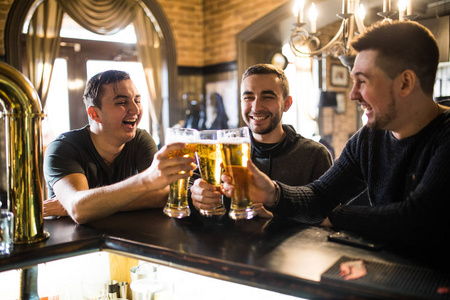 朋友在酒吧。三只快乐的年轻男人在不经意间穿说话，坐在酒吧里喝啤酒一起对抗