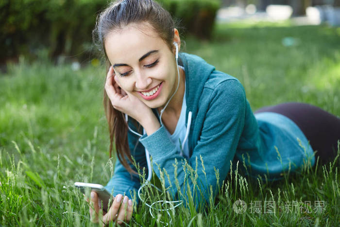 在运动服的年轻和运动女人肖像在于在公园的草地上智能手机