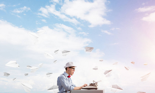 青年作家在帽子和眼镜使用打字机, 而坐在桌子之间的云影像的飞行纸飞机背景