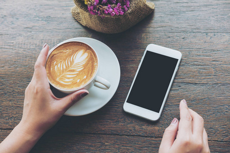 样机形象的白色手机与空白的黑屏和咖啡馆老式木桌上的手热的拿铁咖啡