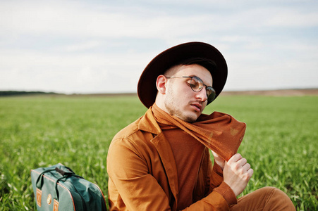 戴眼镜的时尚男士, 棕色夹克和帽子, 绿色田野上摆着袋子