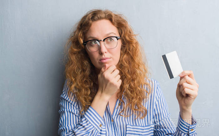 的年轻红头发的女人持有信用卡严肃的面孔思考问题, 非常困惑的想法