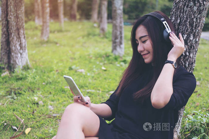一个美丽的亚洲妇女坐在听音乐与耳机和持有智能手机的树木与绿色森林背景