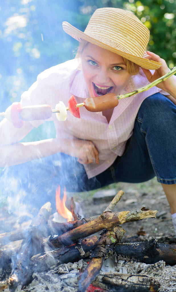 戴草帽的女人试着用棍子咬香肠.女孩吃香肠在棍子.她胃口很大.