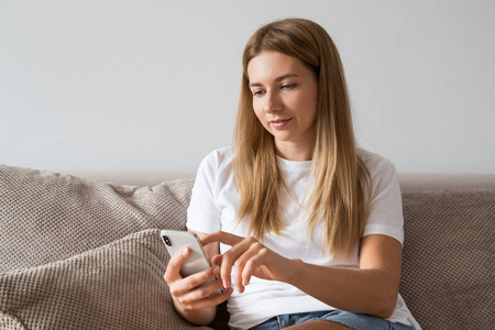 穿着便装的漂亮女孩正用智能手机, 躺在家里的沙发上微笑。妇女在白色 t恤衫浏览互联网