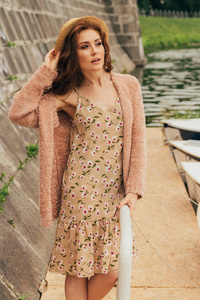 一个美丽的女孩站在码头上, 靠近墙壁和小船。身着花裙, 米色毛衣, 帽子。暑假